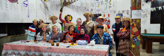 A imagem mostra um grupo animado de pessoas idosas participando de uma festa junina, possivelmente em um centro de convivência ou espaço comunitário. Todos estão vestidos com trajes típicos juninos: chapéus de palha, roupas xadrez, vestidos coloridos, lenços e adereços como tranças falsas e maquiagem no rosto. O ambiente está bem decorado com bandeirinhas, serpentinas coloridas, enfeites de sol, estrelas e elementos típicos das festas de São João. As mesas estão cobertas com toalhas estampadas em estilo tradicional, e há comidas e bebidas típicas sobre elas, como bolo, refrigerante, copos plásticos e pratos descartáveis. No centro da imagem, uma das mulheres ergue uma garrafa em comemoração, acrescentando um ar de celebração ao momento. Ao fundo, vê-se uma churrasqueira grande e, no canto direito, uma mesa de sinuca, o que sugere que o local é usado para outras atividades sociais também. A atmosfera é de alegria, confraternização e celebração da cultura popular brasileira.