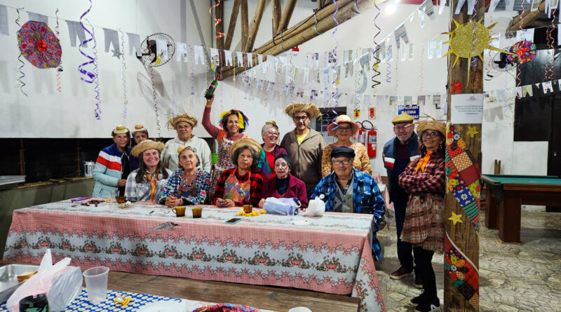 A imagem mostra um grupo animado de pessoas idosas participando de uma festa junina, possivelmente em um centro de convivência ou espaço comunitário. Todos estão vestidos com trajes típicos juninos: chapéus de palha, roupas xadrez, vestidos coloridos, lenços e adereços como tranças falsas e maquiagem no rosto. O ambiente está bem decorado com bandeirinhas, serpentinas coloridas, enfeites de sol, estrelas e elementos típicos das festas de São João. As mesas estão cobertas com toalhas estampadas em estilo tradicional, e há comidas e bebidas típicas sobre elas, como bolo, refrigerante, copos plásticos e pratos descartáveis. No centro da imagem, uma das mulheres ergue uma garrafa em comemoração, acrescentando um ar de celebração ao momento. Ao fundo, vê-se uma churrasqueira grande e, no canto direito, uma mesa de sinuca, o que sugere que o local é usado para outras atividades sociais também. A atmosfera é de alegria, confraternização e celebração da cultura popular brasileira.