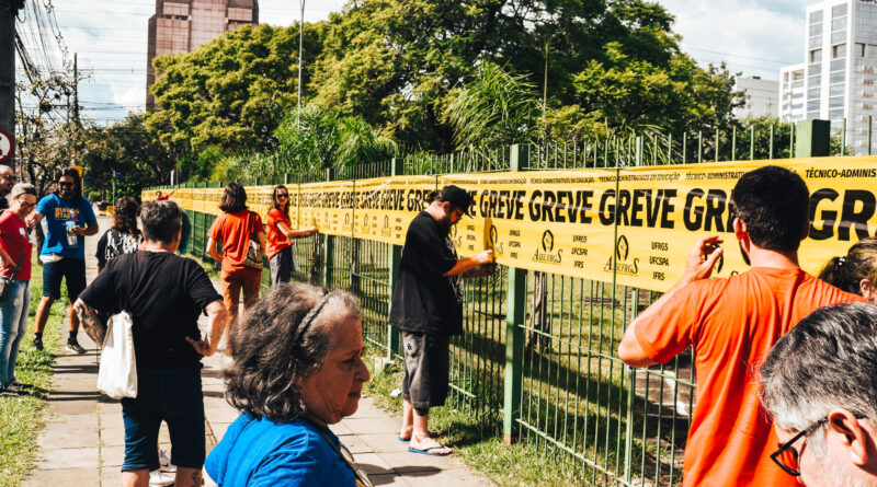 A imagem mostra um grupo de pessoas reunidas ao longo de uma calçada, em frente a uma cerca de metal pintada de verde. É um dia ensolarado, com árvores grandes e frondosas ao fundo, além de prédios urbanos visíveis mais atrás. As pessoas, de diferentes idades e gêneros, estão espalhadas ao longo da calçada e próximas à cerca. Algumas vestem camisetas vermelhas e outras roupas casuais. Elas parecem envolvidas em uma atividade coletiva: várias estão fixando uma longa faixa amarela na cerca. A faixa se estende por grande parte do comprimento da grade e contém repetidamente a palavra “GREVE” em letras grandes e pretas, além de outros textos menores e logotipos. No primeiro plano, à direita, há um homem com cabelo curto e barba grisalha, usando óculos e olhando para baixo. Mais ao centro, uma mulher de cabelo curto e grisalho, vestindo uma camiseta azul, observa a cena de lado. Ao longo da cerca, outras pessoas estão colando ou ajustando a faixa, algumas com as mãos levantadas, concentradas na tarefa. O ambiente sugere um contexto de mobilização ou protesto, com clima colaborativo e organizado, em uma área urbana arborizada.