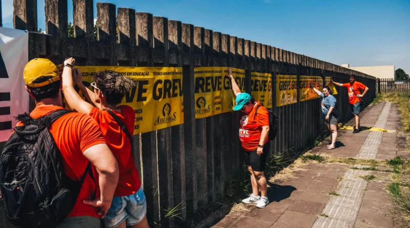 A imagem mostra um grupo de pessoas em uma ação ao ar livre, em um dia ensolarado, com céu azul e sem nuvens. Elas estão posicionadas ao longo de uma calçada, ao lado de uma longa grade de concreto com pilares verticais, que forma uma espécie de muro vazado. As pessoas vestem camisetas vermelhas e parecem estar colando ou ajustando cartazes amarelos na grade. Esses cartazes estão alinhados lado a lado e trazem a palavra “GREVE” em letras grandes e escuras, indicando uma mobilização ou protesto. Algumas pessoas usam bonés e mochilas, e estão concentradas na tarefa de fixar os cartazes, com os braços levantados em direção à grade. A cena é iluminada por luz forte do sol, que cria sombras marcadas no chão e na estrutura de concreto. A calçada é feita de placas retangulares, com uma faixa tátil no centro, indicando acessibilidade para pessoas com deficiência visual. Ao fundo, há mais pessoas participando da mesma atividade, além de uma área com vegetação baixa e um prédio claro, sugerindo que o local pode ser um campus ou espaço institucional.