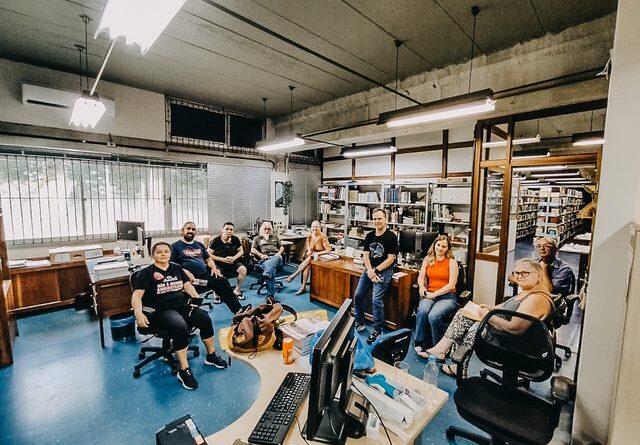 A imagem mostra um grupo de cerca de dez pessoas reunidas em uma sala de trabalho que lembra um escritório ou laboratório. O ambiente é interno, com iluminação artificial vinda de luminárias retangulares no teto. O piso é azul e há mesas de madeira com computadores, teclados e outros equipamentos espalhados pelo espaço. As pessoas estão distribuídas em cadeiras giratórias e algumas sentadas sobre as mesas, formando um semicírculo voltado para a câmera. Elas são de diferentes idades e gêneros, vestem roupas casuais — como camisetas, calças e tênis — e aparentam estar em um momento de pausa ou reunião informal. Algumas sorriem ou olham diretamente para a câmera, enquanto outras mantêm uma postura mais relaxada. Ao fundo, há estantes cheias de documentos, livros ou caixas organizadas, além de uma janela com grades e persianas parcialmente abertas, deixando entrar um pouco de luz externa. O ambiente sugere um local de trabalho coletivo, possivelmente em uma instituição ou setor administrativo, com clima de convivência e colaboração entre os presentes.