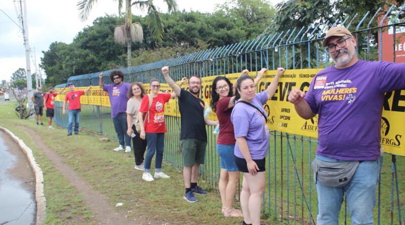 A imagem mostra um grupo de pessoas alinhadas ao longo de uma grade metálica azul, em uma área externa, próxima a uma rua. O dia está claro, mas o céu parece levemente nublado. Ao longo da grade, estão fixados vários cartazes amarelos, colocados lado a lado, formando uma faixa contínua. Esses cartazes trazem a palavra “GREVE” em destaque, indicando uma mobilização coletiva. As pessoas estão posicionadas em fila, espaçadas umas das outras, todas voltadas para a câmera. Muitas delas levantam um dos braços, com o punho fechado, em um gesto associado à luta e resistência. O grupo é diverso, composto por homens e mulheres de diferentes idades. Algumas pessoas usam óculos, e várias vestem camisetas em cores como vermelho e roxo, algumas com dizeres relacionados a causas sociais e sindicais. Em primeiro plano, à direita da imagem, há um homem usando camiseta roxa, boné e óculos, com barba e expressão séria, também com o braço erguido. Ao longo da fila, outras pessoas sorriem ou mantêm expressões firmes, transmitindo união e engajamento. O ambiente ao redor inclui uma faixa de grama entre a calçada e a rua, postes de energia e árvores ao fundo, incluindo uma palmeira. A cena como um todo transmite a ideia de organização coletiva, solidariedade e participação em um movimento de greve, com o grupo demonstrando apoio à causa de forma visível e coordenada.