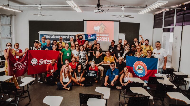 Foto de um grupo grande de pessoas reunidas em uma sala de aula ou auditório. Ao todo, cerca de 30 a 40 pessoas, de diferentes idades e gêneros, posam juntas sorrindo para a câmera. A maioria está em pé ao fundo, enquanto algumas estão sentadas ou agachadas na frente. O ambiente possui cadeiras escolares com mesas acopladas espalhadas pelo espaço, ventiladores no teto e iluminação fluorescente. Ao fundo, há um telão de projeção exibindo um slide com um punho cerrado em fundo vermelho e a frase “Encontro Internacional Antifascista”. Também há uma faixa estendida na parede com dizeres relacionados a uma organização sindical. Algumas pessoas seguram bandeiras e faixas vermelhas e azuis com logotipos de sindicatos e entidades, reforçando o caráter político e coletivo do encontro. No centro da imagem, uma pessoa segura um cartaz com a palavra “GREVE”. A cena transmite um clima de união, mobilização e engajamento político, com todos reunidos de forma organizada para registrar o momento coletivo.