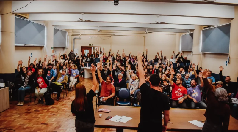 A imagem mostra uma sala ampla de reunião, com piso de madeira e iluminação clara, provavelmente em um prédio institucional ou universitário. O ambiente tem fileiras de cadeiras ocupadas por dezenas de pessoas, de diferentes idades e aparências, formando uma plateia bastante cheia. Na parte da frente da sala, há uma mesa comprida com alguns papéis e objetos sobre ela. De costas para quem observa a imagem, três pessoas estão em pé atrás dessa mesa, voltadas para o público. Elas levantam um dos braços, como se estivessem conduzindo uma votação ou pedindo manifestação dos presentes. A grande maioria das pessoas sentadas também está com um braço levantado, acompanhando o gesto, o que sugere um momento coletivo de decisão, como uma assembleia. As expressões aparentam ser atentas e engajadas. Ao fundo, é possível ver portas, paredes claras, janelas com cortinas e alguns equipamentos como ventiladores de teto. O ambiente transmite a sensação de um encontro organizado, participativo e com forte envolvimento do grupo presente.
