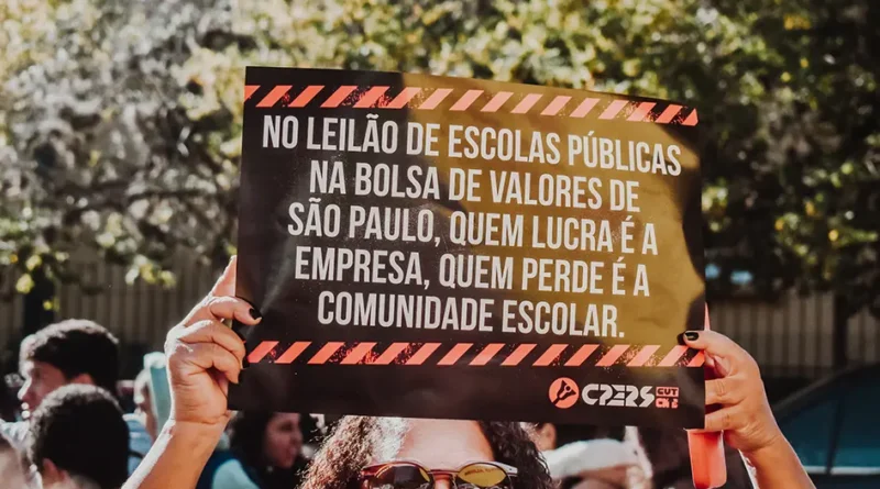 Fotografia colorida registrada em um ato público ao ar livre, durante o dia. Em primeiro plano, uma mulher de cabelos cacheados escuros e óculos escuros levanta acima da cabeça um cartaz preto com letras brancas em destaque. O cartaz possui faixas diagonais em vermelho e preto nas bordas superior e inferior, lembrando sinalização de alerta. O texto do cartaz diz: “No leilão de escolas públicas na Bolsa de Valores de São Paulo, quem lucra é a empresa, quem perde é a comunidade escolar.” No canto inferior direito aparece o logotipo do CPERS e da CUT. A mulher aparece apenas parcialmente, com o rosto coberto em parte pelo cartaz. Ao fundo, desfocado, há outras pessoas participando da manifestação e árvores iluminadas pela luz do sol, criando um clima de mobilização social e protesto em defesa da educação pública.
