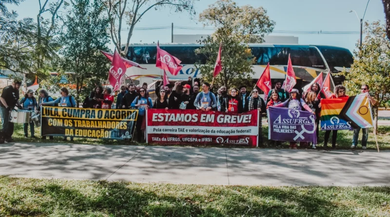 Fotografia horizontal registrada ao ar livre durante o dia mostra um grupo numeroso de manifestantes reunidos em uma calçada arborizada. Ao fundo, há um ônibus branco estacionado e árvores altas com galhos espalhados pelo céu claro. As pessoas estão lado a lado segurando bandeiras vermelhas e faixas de greve ligadas à Assufrgs e aos trabalhadores da educação federal. No centro da imagem, uma grande faixa vermelha traz a frase em letras brancas: “Estamos em greve!”, acompanhada de um texto menor sobre a valorização da carreira dos técnicos-administrativos em educação. À esquerda, outra faixa preta e amarela diz: “Lula, cumpra o acordo com os trabalhadores da educação”. À direita, um banner roxo destaca uma campanha “pela vida das mulheres”, e ao lado há uma bandeira com cores do movimento LGBTQIA+. O grupo é diverso, composto por homens e mulheres de diferentes idades, muitos usando camisetas, mochilas, óculos escuros e roupas casuais. Algumas pessoas levantam bandeiras enquanto outras seguram instrumentos de percussão. A cena transmite clima de mobilização coletiva, protesto e organização sindical.